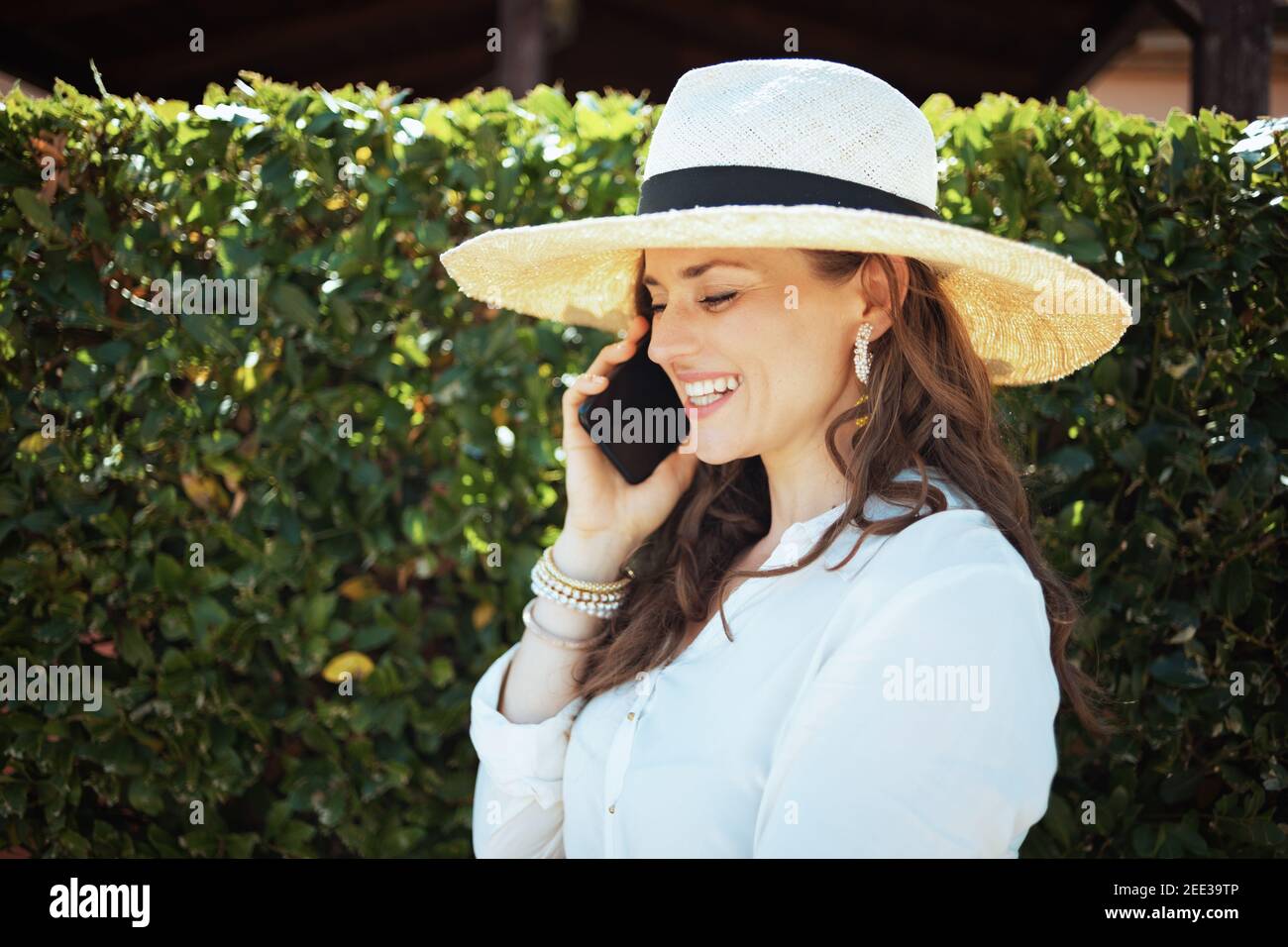 sorridente casalinga di mezza età in camicia bianca con cappello che parla su uno smartphone all'aperto vicino al muro verde. Foto Stock