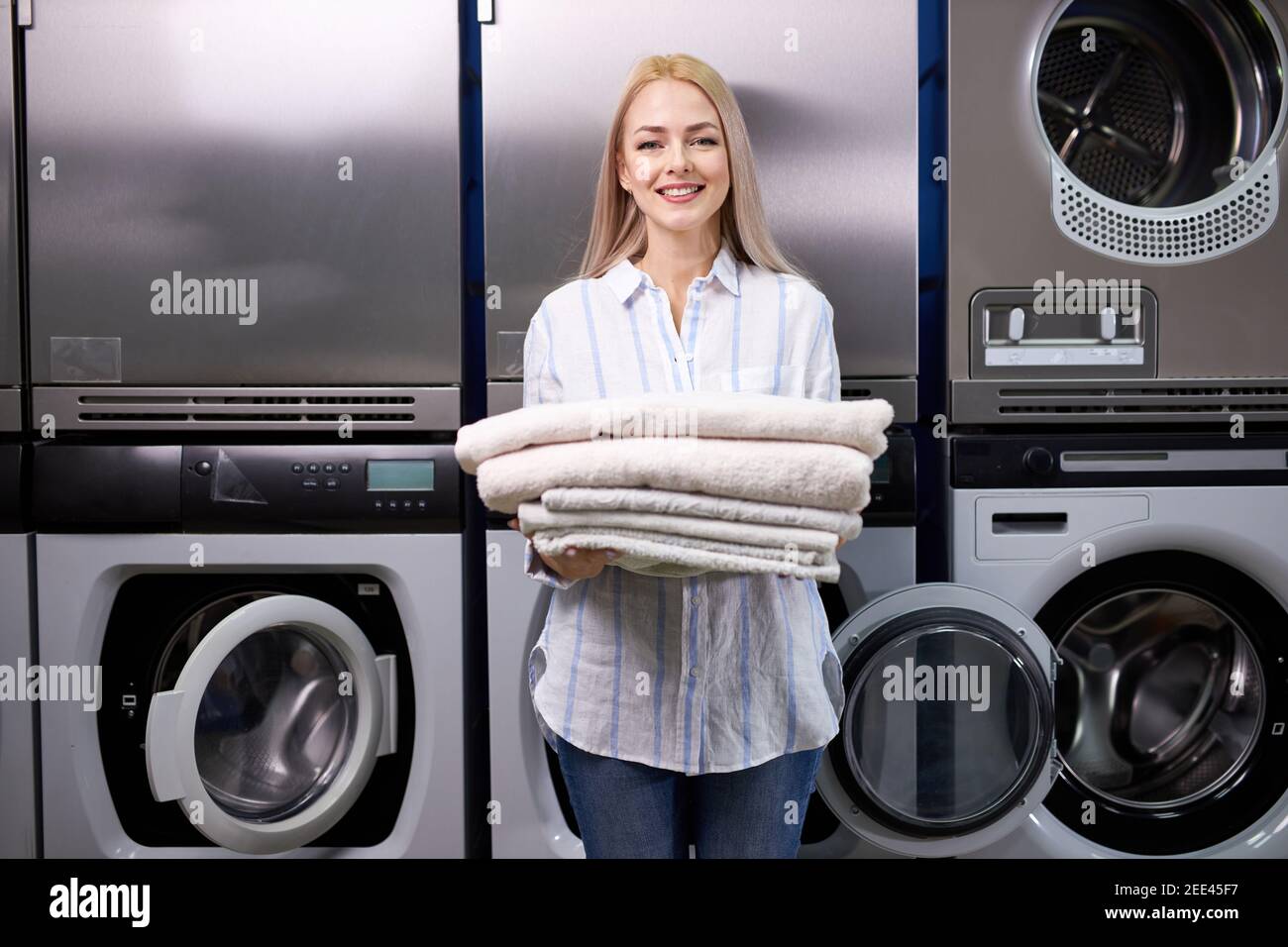 donna bionda sorridente felice di fare i chores alla lavanderia, tenendo una pila di asciugamani puliti freschi. un sacco di lavatrici in background Foto Stock donna bionda sorridente felice di fare i chores alla lavanderia, tenendo una pila di asciugamani puliti freschi. un sacco di lavatrici in background Foto Stock