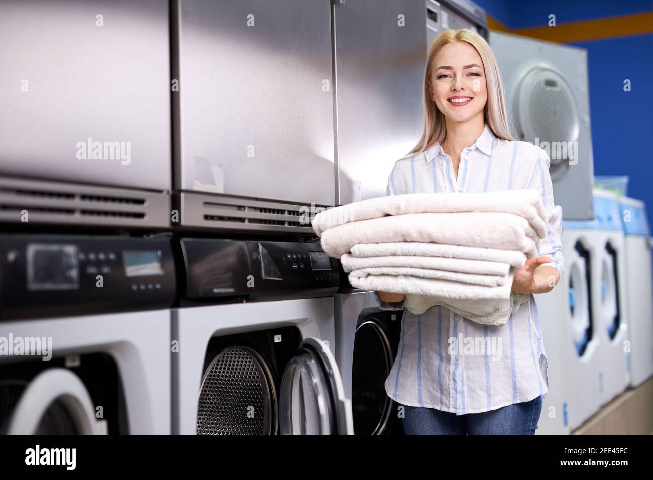 donna bionda sorridente felice di fare i chores alla lavanderia, tenendo una pila di asciugamani puliti freschi. un sacco di lavatrici in background Foto Stock donna bionda sorridente felice di fare i chores alla lavanderia, tenendo una pila di asciugamani puliti freschi. un sacco di lavatrici in background Foto Stock