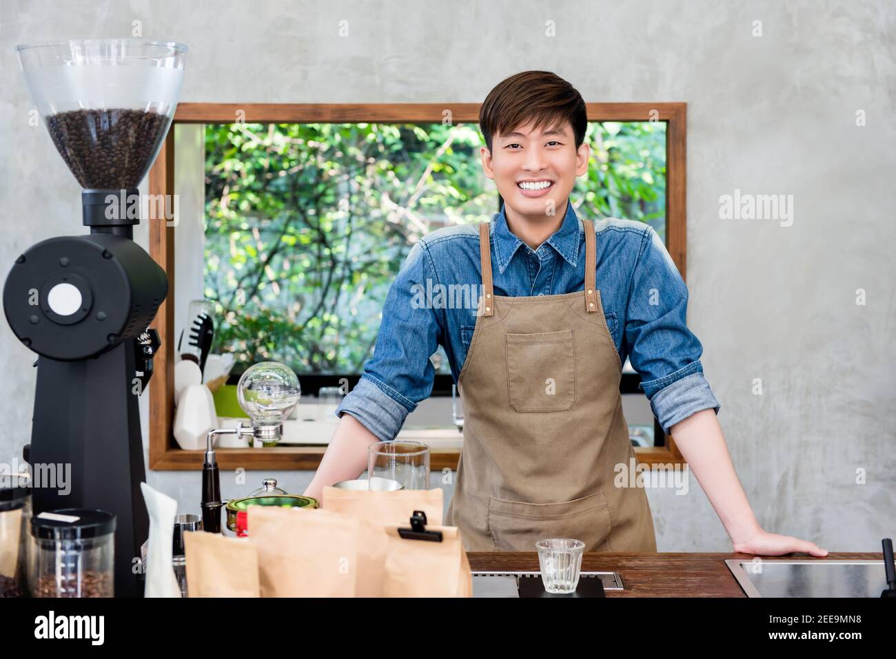 Allegro e sorridente giovane imprenditore asiatico in piedi presso la caffetteria sportello pronto ad accogliere e fornire assistenza al cliente Foto Stock