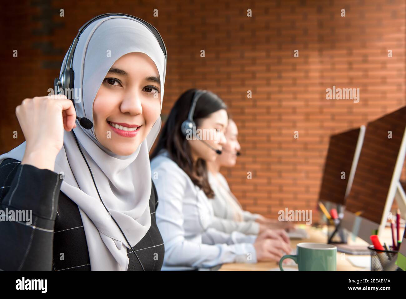 Sorridente bella donna d'affari musulmana agente di telemarketing o operatore di servizio di cliente lavorare in azienda con il suo team in un call center Foto Stock