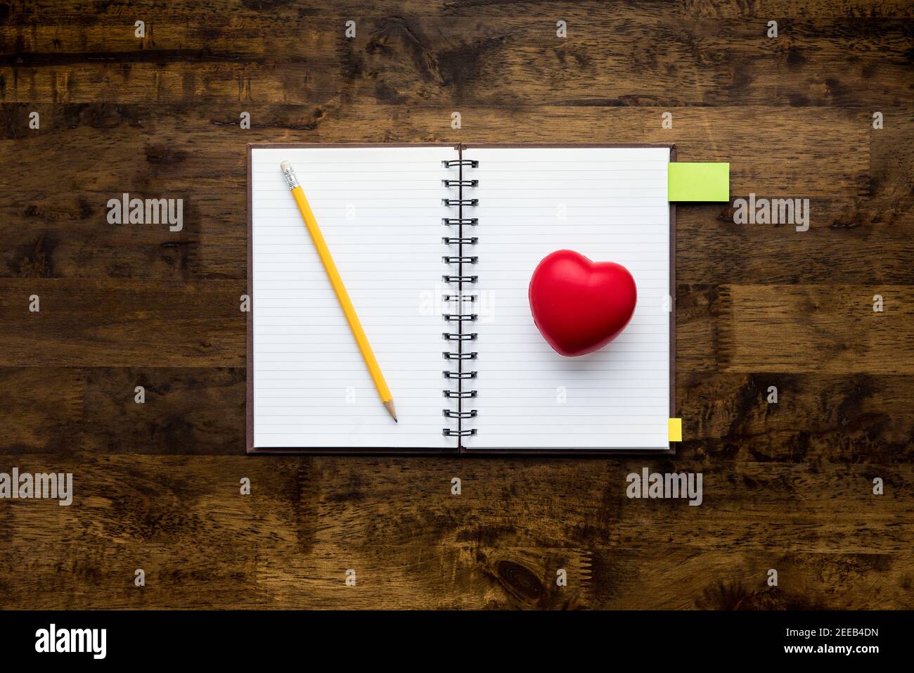 Taccuino vuoto aperto con matita e pallina a forma di cuore rosso sullo sfondo del tavolo in legno, vista dall'alto Foto Stock