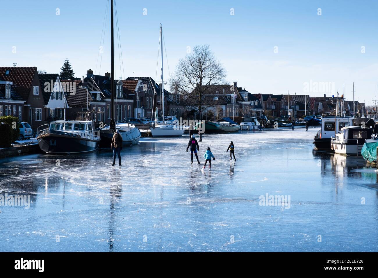 La gente pattina sul ghiaccio di Het Dok a Lemmer con le navi di destra e di sinistra congelate nel ghiaccio sulla banchina con le case. Scenario invernale soleggiato Foto Stock