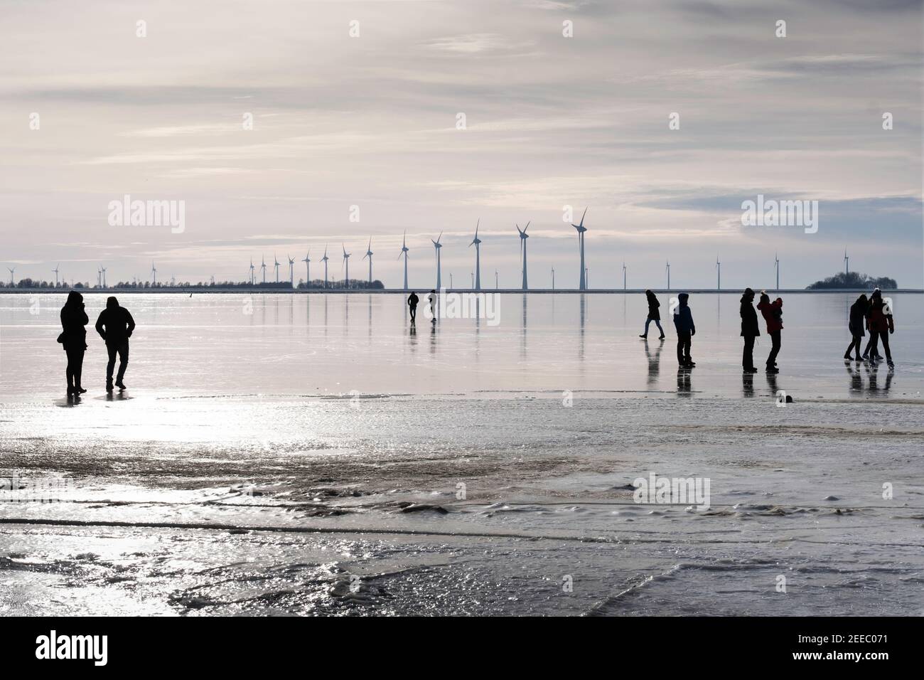 Silhouette di persone sul ghiaccio della baia ghiacciata di Lemmer, Paesi Bassi. In background mulini a vento per la generazione di energia Foto Stock