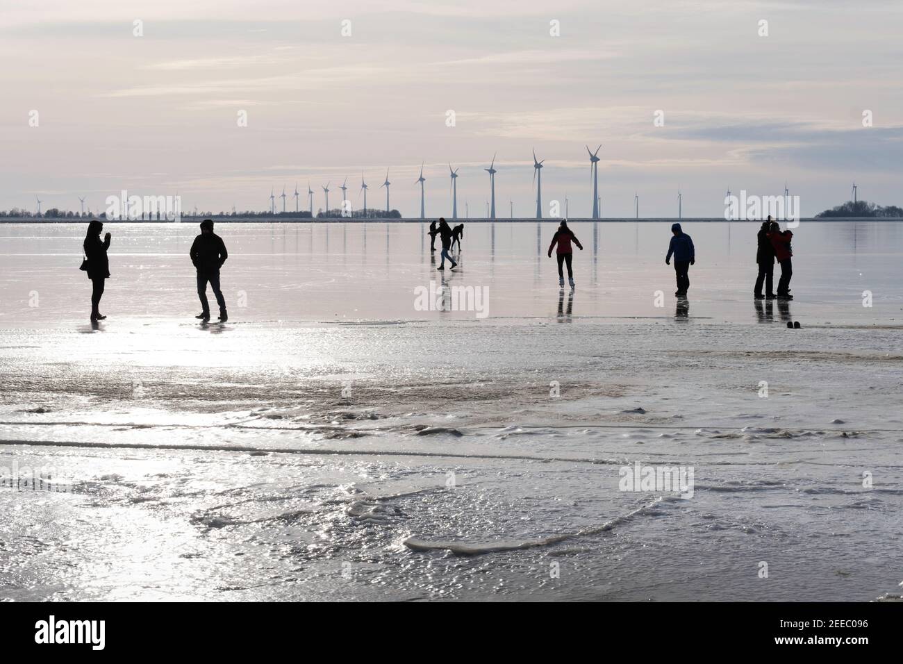 Silhouette di persone sul ghiaccio della baia ghiacciata di Lemmer, Paesi Bassi. In background mulini a vento per la generazione di energia Foto Stock