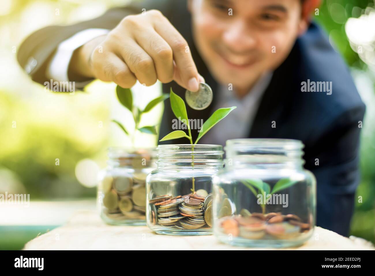 Uomo d'affari che mette la moneta nel vaso di vetro con una pianta giovane, dimostrando la crescita finanziaria attraverso piani di risparmio e piani di investimento Foto Stock