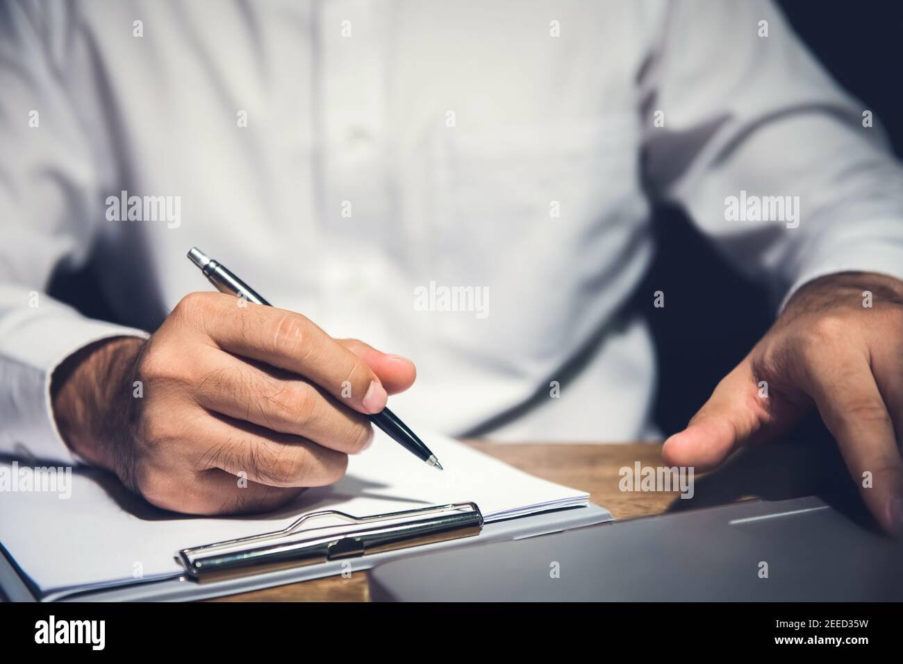 Uomo d'affari che lavora straordinario alla sua scrivania in ufficio a tarda notte, utilizzando un computer portatile e che sta per scrivere su carta Foto Stock