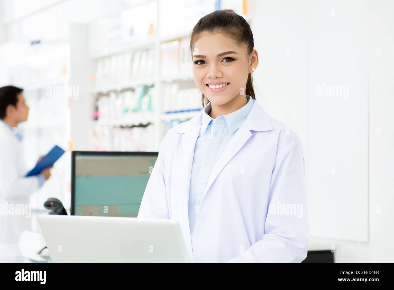 Giovane donna asiatica sorridente farmacista in abito bianco uniforme lavoro su computer portatile in farmacia o negozio di farmacia Foto Stock