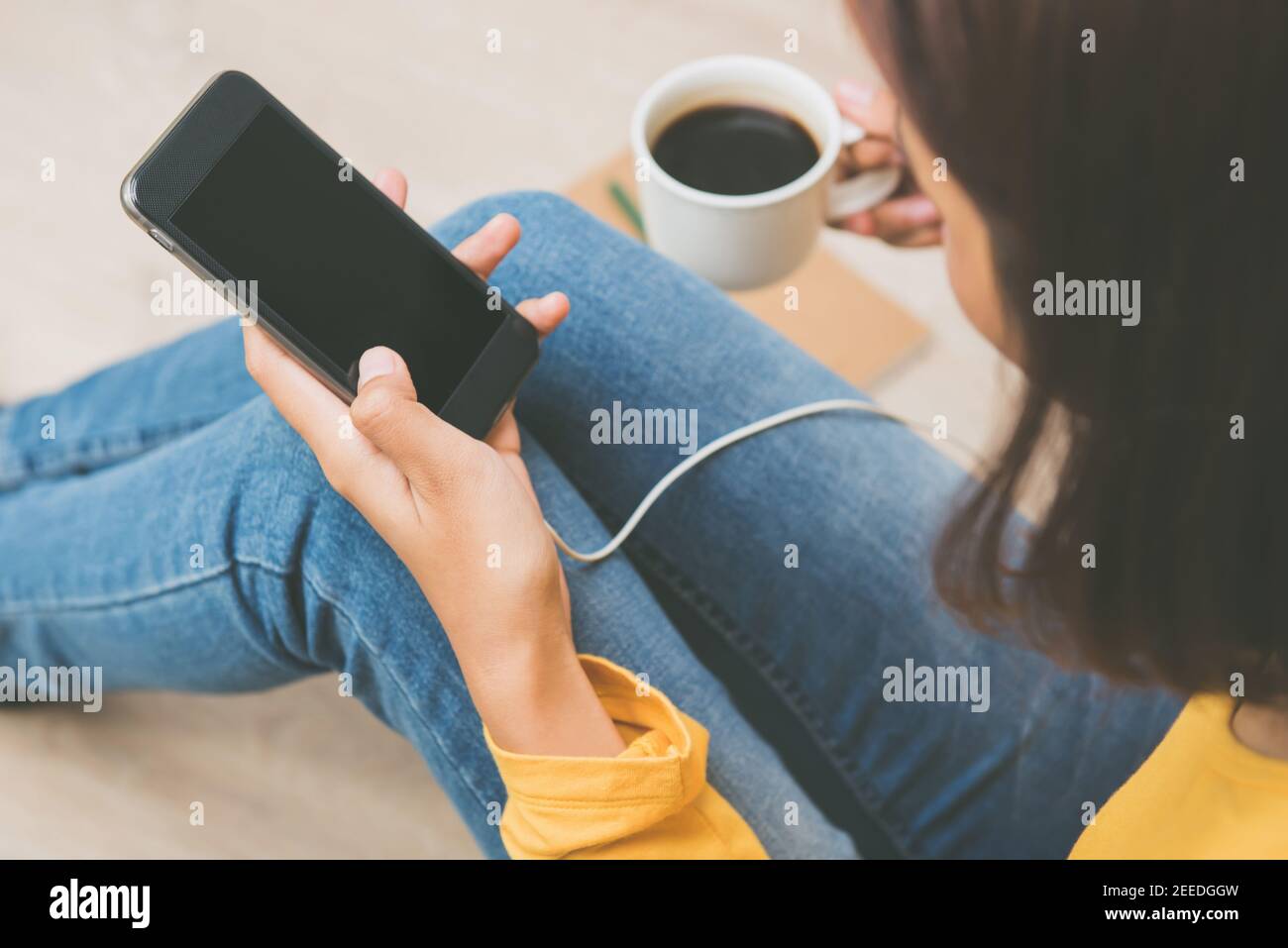 Donna informale rilassante seduta a casa bere caffè e utilizzare smartphone sul pavimento Foto Stock