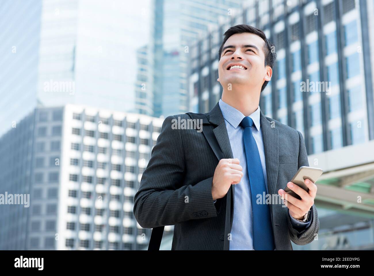 Giovane felice uomo d'affari sorridente che guarda in su mentre tiene il telefono mobile e borsa da trasporto in città - viaggio d'affari e. concetti relativi al roaming mobile Foto Stock