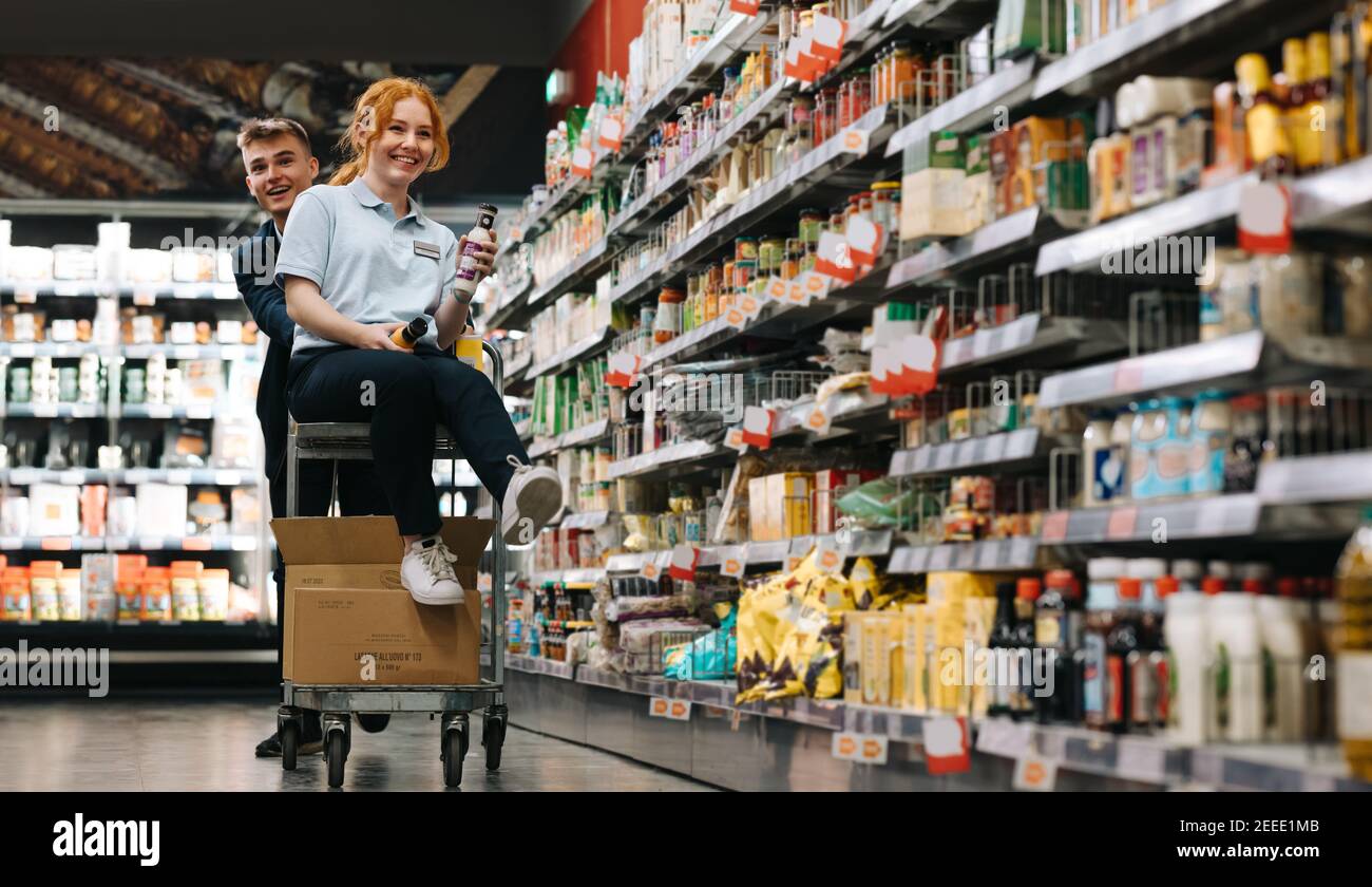 Due giovani lavoratori che hanno merci di imballaggio divertenti. Donna seduta sul carrello con un collega uomo che spinge nella corsia del supermercato. Foto Stock