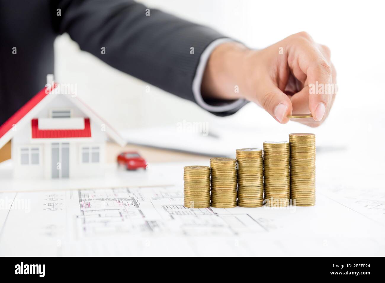 Uomo d'affari che mette la moneta sulla cima di pile di denaro con sfocato modello di casa in background - immobiliare finanziario e investimenti concetti Foto Stock