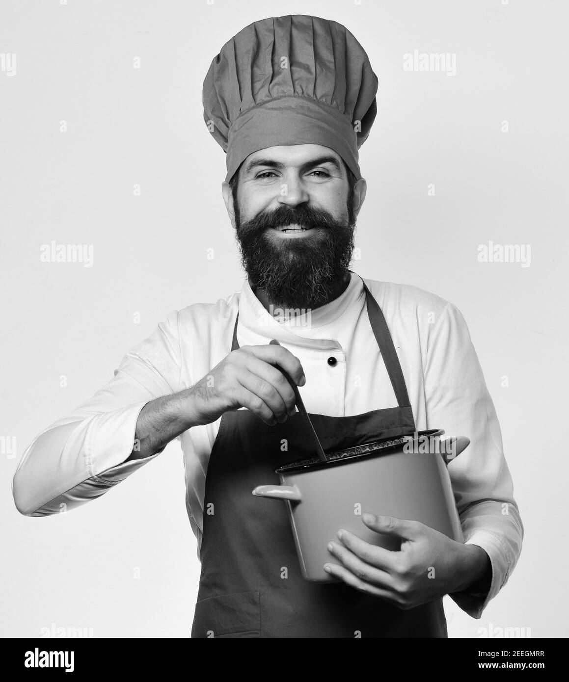 Ristorante cucina concetto. Lo chef mescola zuppa o composta. L'uomo con la barba tiene il vaso rosso su sfondo bianco. Cuoco con viso felice in uniforme di borgogna ha casseruola con mestolo. Foto Stock
