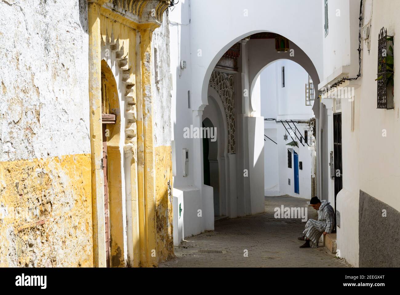 Un uomo che indossa una djellaba a righe si trova di fronte a una piccola moschea, sulla soglia di una casa nella medina di Tangeri, Marocco. Foto Stock