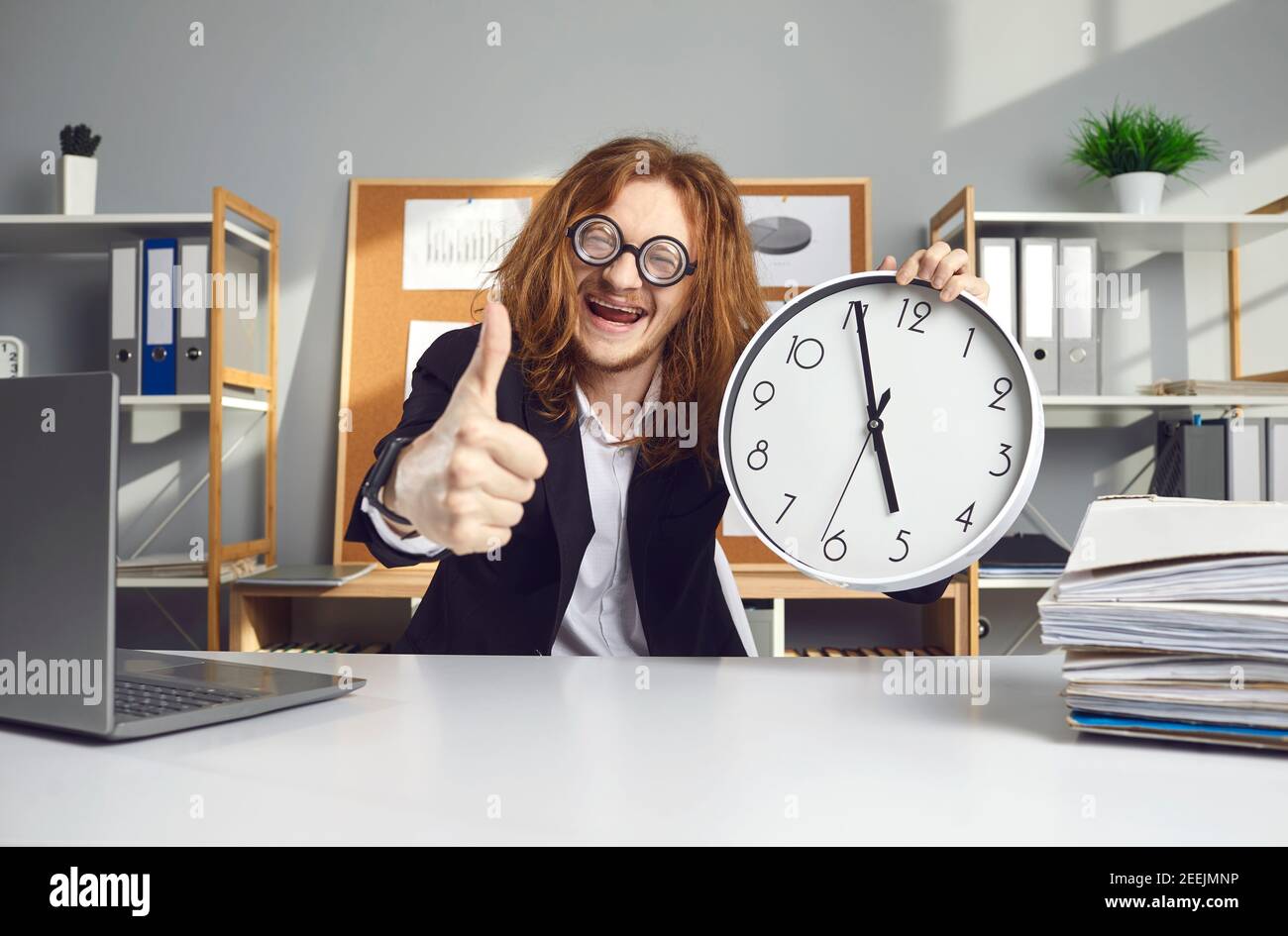 Un buon lavoratore seduto alla scrivania, tenendo l'orologio che dice che è alle 17:00 e dando i pollici Foto Stock