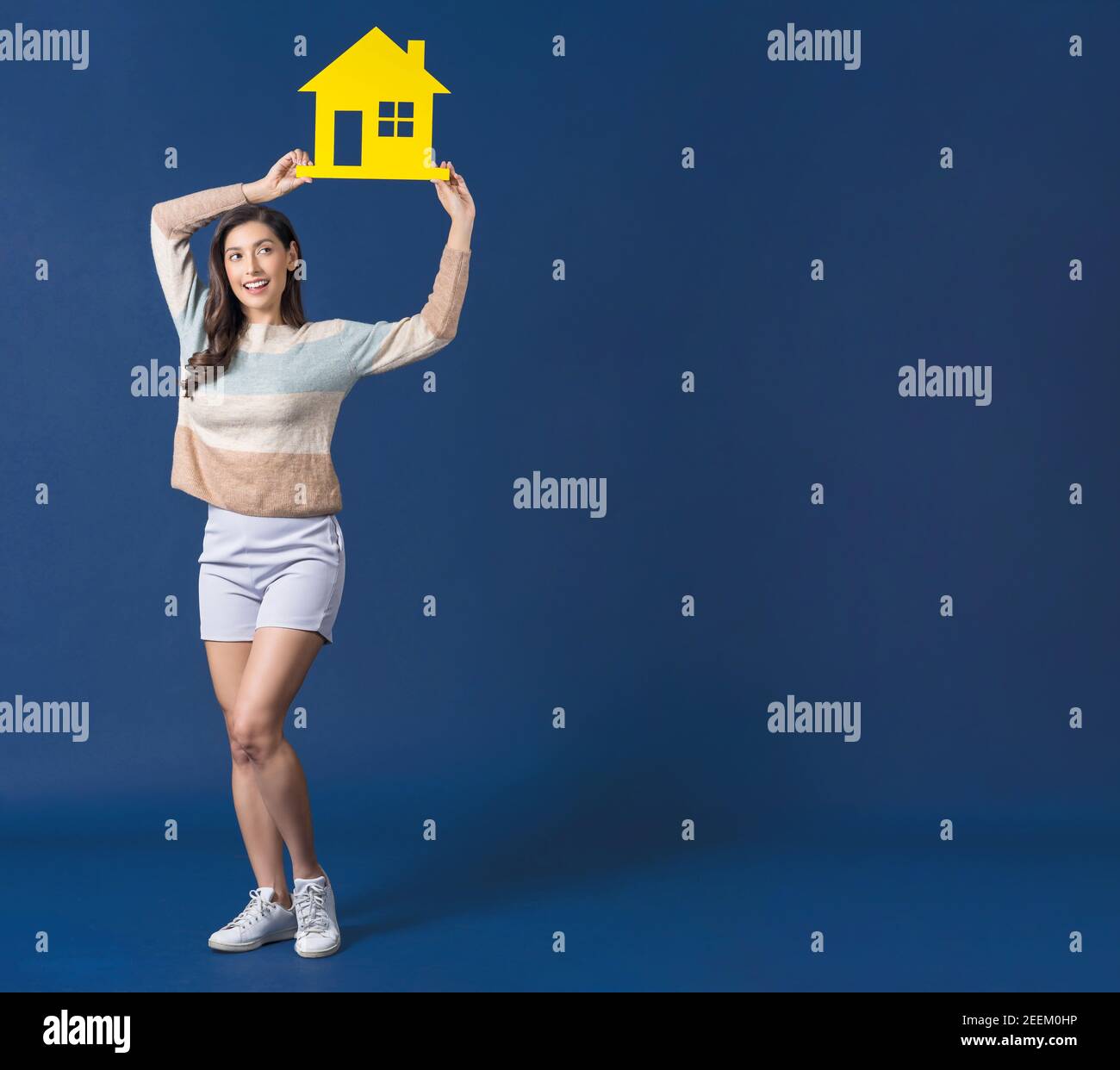 Felice giovane donna asiatica sorridente che tiene e mostra giallo colore casa mockup carta su sfondo blu colore, pensando per gli investimenti, le vendite e insur Foto Stock Felice giovane donna asiatica sorridente che tiene e mostra giallo colore casa mockup carta su sfondo blu colore, pensando per gli investimenti, le vendite e insur Foto Stock