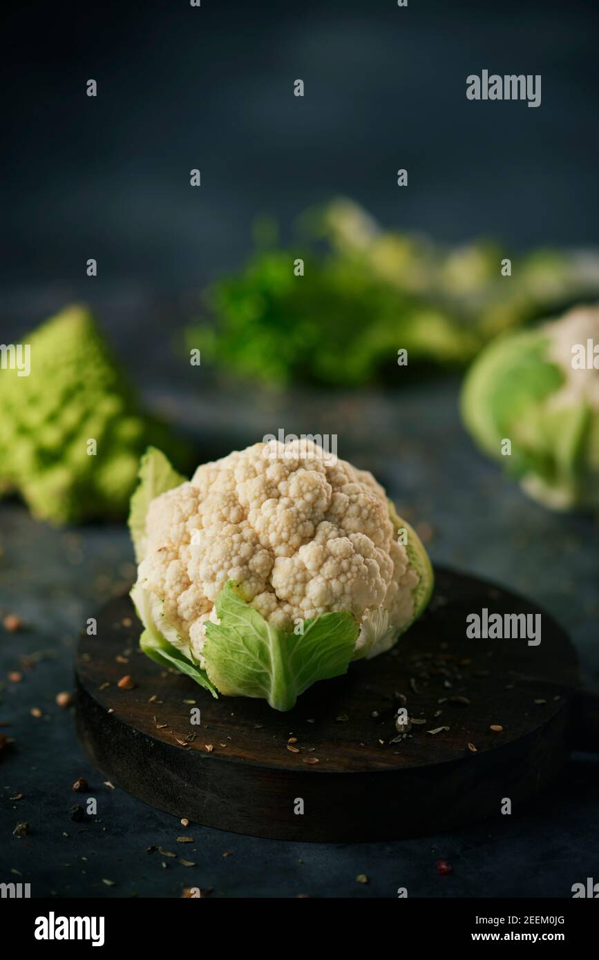 una testa cruda del cavolfiore del bambino in una piastra di legno, posta su una superficie scura della pietra spruzzata con semi e spezie differenti e un broccoli del romanesco del bambino Foto Stock