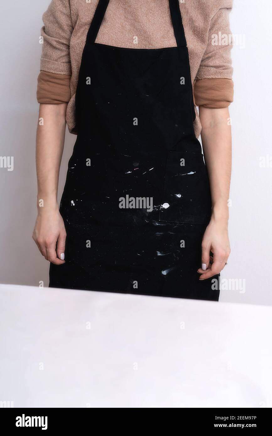 Una donna artista in un grembiule nero con gocce di vernice si trova contro il muro della sua officina. Sfondo artistico con posto per il testo. Foto Stock