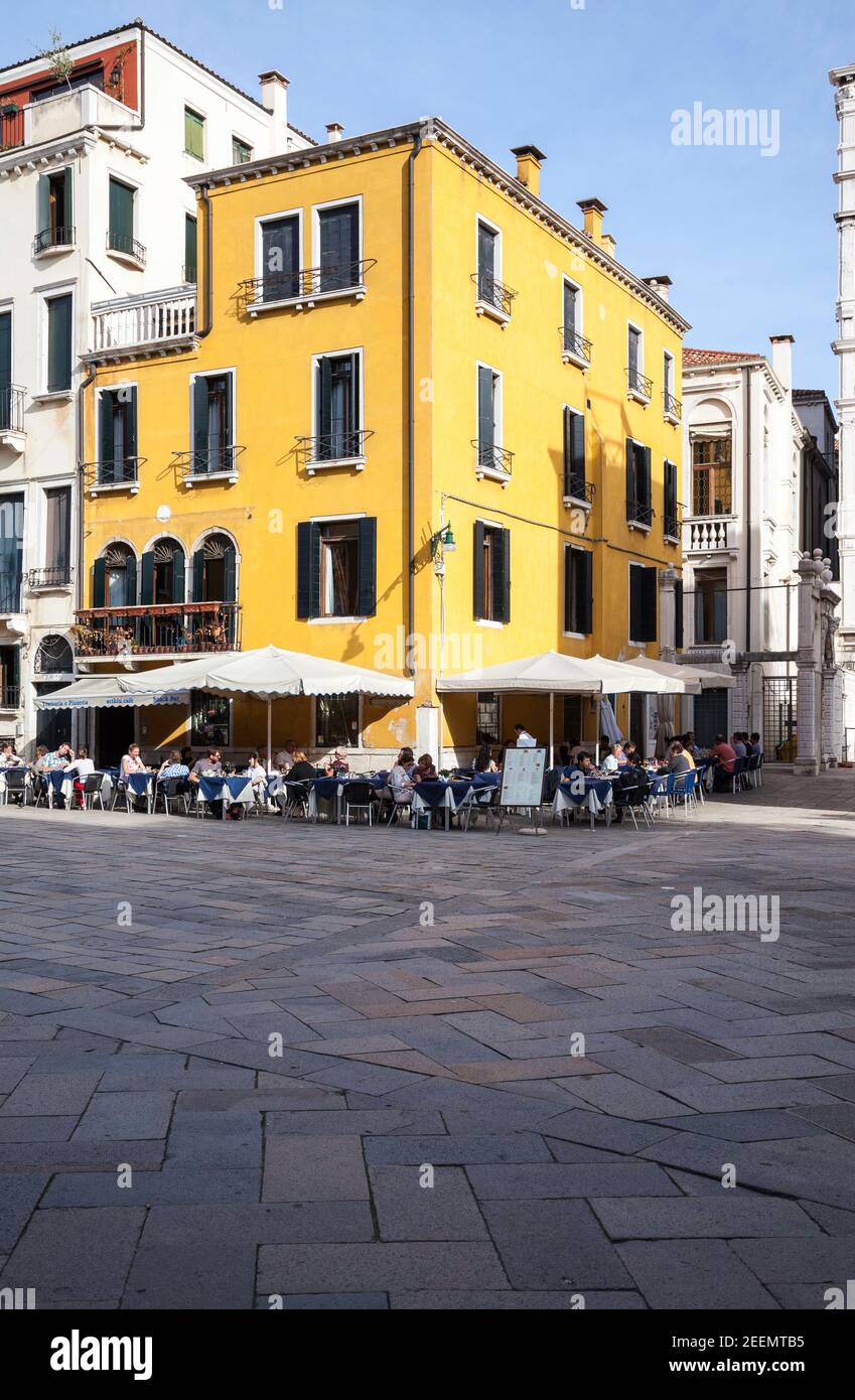Le persone si rilassano al sole sotto un cielo blu limpido in un caffè con pavimento dipinto di giallo luminoso su campo Santo Stefano a Venezia, Italia con spazio per le copie Foto Stock