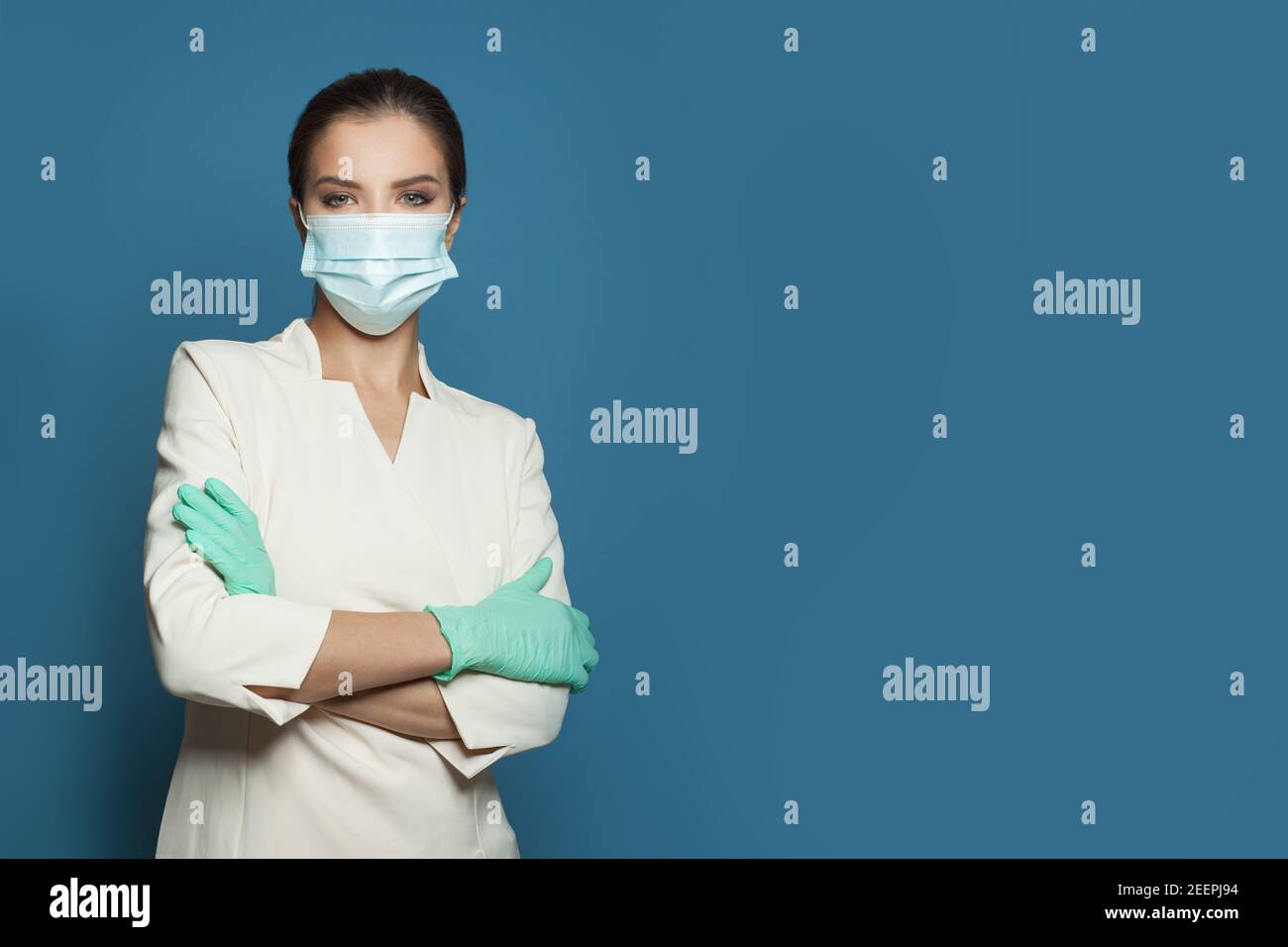 Donna medico sicura che indossa una maschera medica protettiva e guanti chirurgici braccia in piedi incrociate su sfondo blu. Medicina, sicurezza e virus covid- Foto Stock