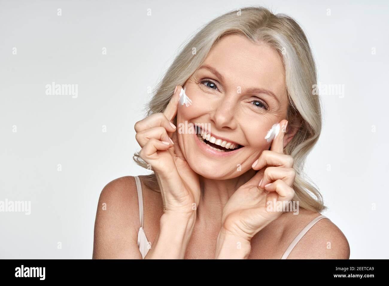 Donna felice di mezza età che applica crema sulla pelle del viso isolato su sfondo bianco. Foto Stock