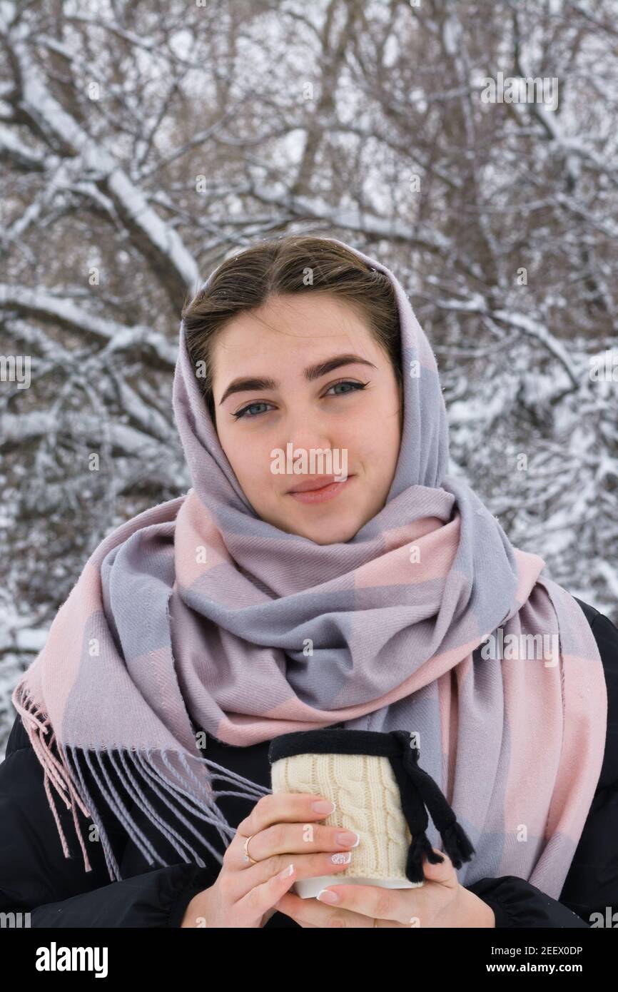 Ritratto di bella giovane donna con occhi blu. Passeggiata invernale nella foresta innevata. Tazza con bevanda calda in mano. Foto Stock