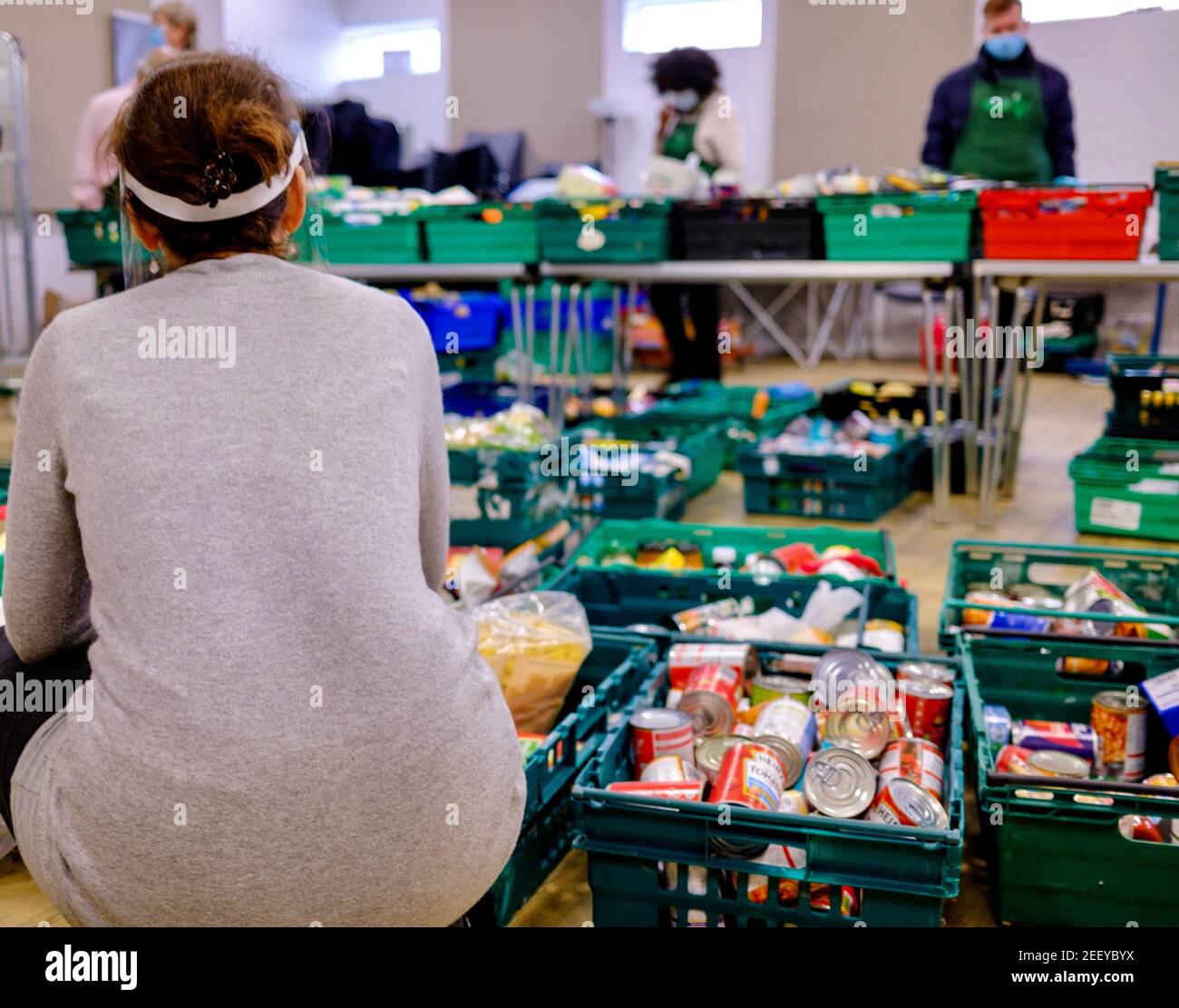 Il volontario della banca di cibo a Trussell Trust foodbank accoccolato accanto a casse di cibo donato con altri volontari cernendo e raccogliendo cibo. Londra, Regno Unito Foto Stock