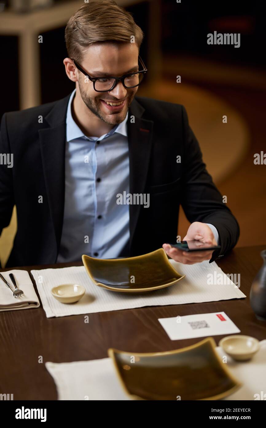 Gentiluomo soddisfatto dai capelli giusti in abbigliamento formale sorridente durante la scansione del codice QR di un menu del ristorante Foto Stock