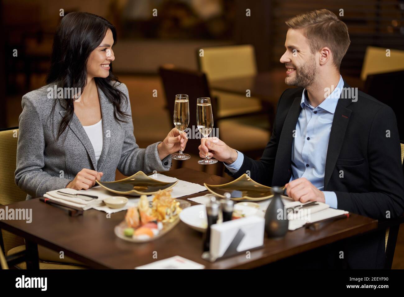 Bella giovane coppia sorridente mentre si tengono bicchieri di champagne e si siede al tavolo servito in una caffetteria Foto Stock