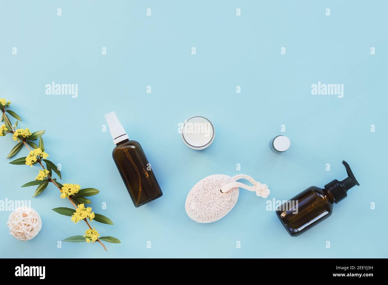 Cosmetici naturali, crema, olio essenziale, tonico della pelle, pietra pomice e sprig con fiori gialli su un tavolo blu. Concetto di bellezza, spa e benessere. In alto Foto Stock