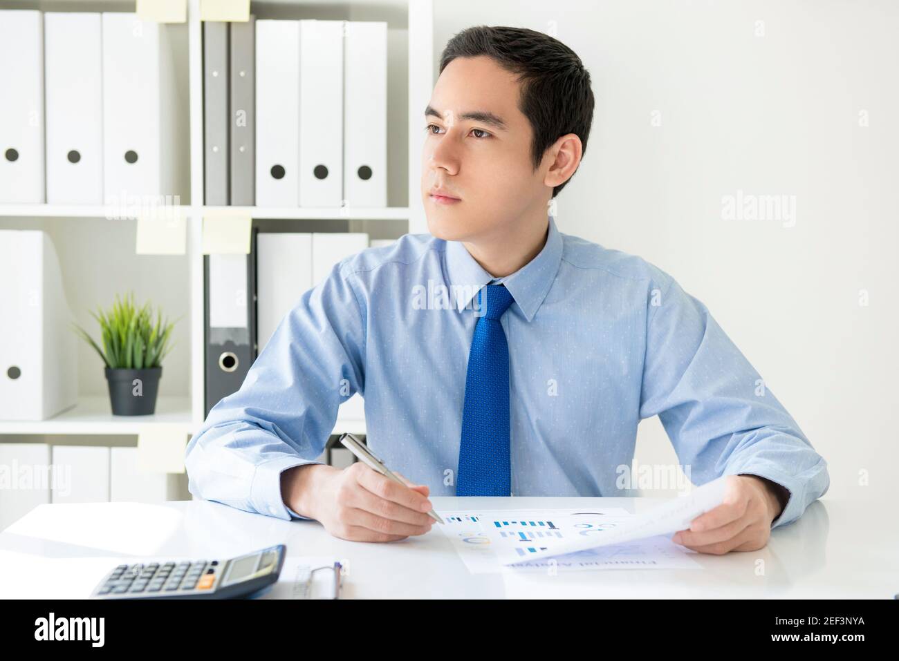 Giovane uomo d'affari asiatico che guarda fuori e pensa mentre si siede scrivania in ufficio Foto Stock