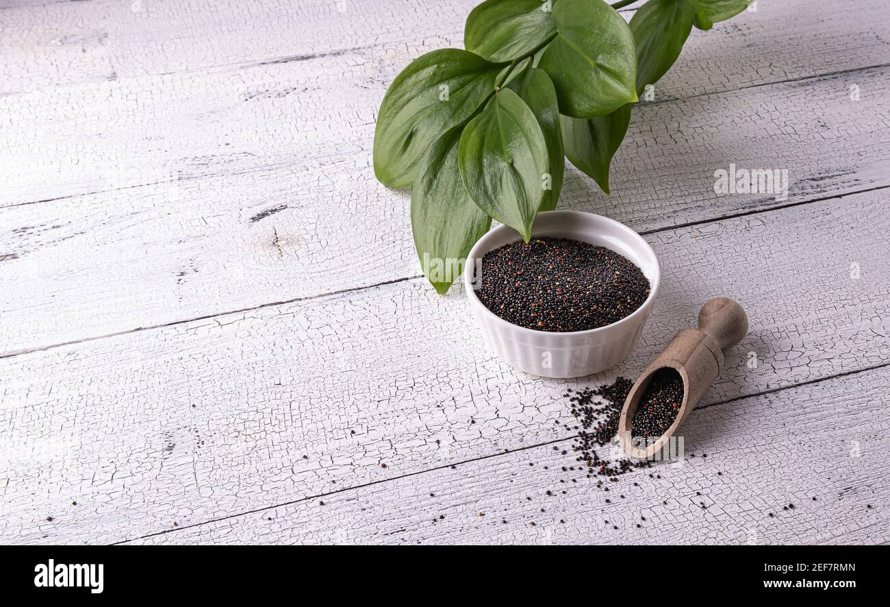 Semi neri di quinoa peruviana crudi in ciotola bianca e paletta di legno con semi su tavola di legno bianco. Pochi semi sono sparsi intorno. Foglie verdi sul dorso Foto Stock