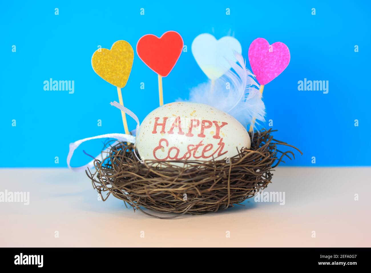 Uovo di Pasqua con Pasqua felice inscritto in un nido, cuori decorativi multicolore e piuma bianca su uno sfondo blu luminoso. Foto Stock