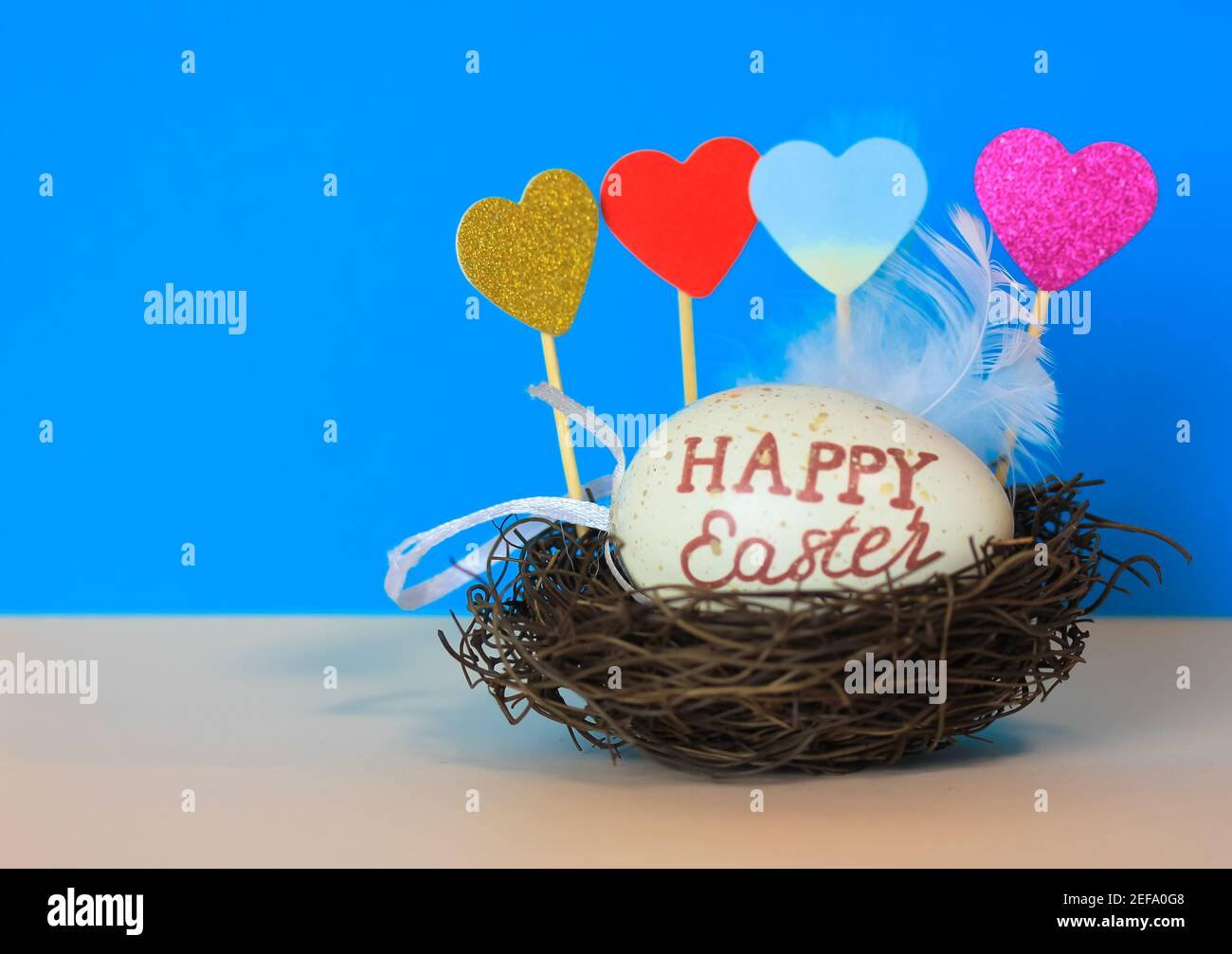 Uovo di Pasqua con Pasqua felice inscritto in un nido, cuori decorativi multicolore e piuma bianca su uno sfondo blu luminoso. Foto Stock