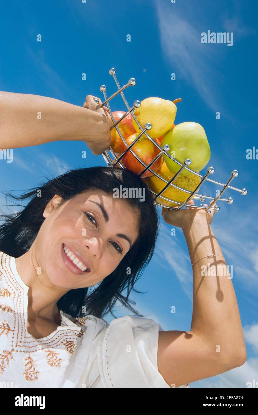 Vista ad angolo basso di una donna adulta media che tiene un vassoio di frutta sulla testa e sorridente Foto Stock