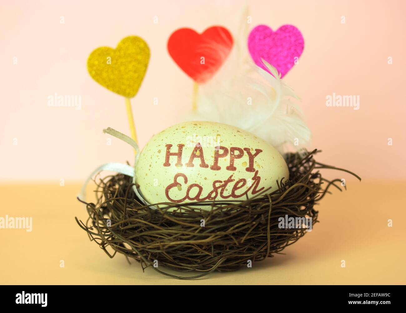 Uovo di Pasqua con Pasqua felice inscritto in un nido, cuori decorativi multicolore e una piuma bianca. Sfondo color pesca chiaro e caldo. Foto Stock
