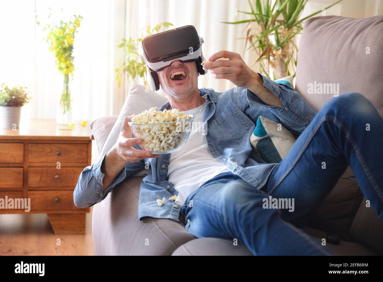 Uomo che guarda contenuti multimediali in occhiali di realtà virtuale mangiare popcorn sdraiati sul divano di casa Foto Stock