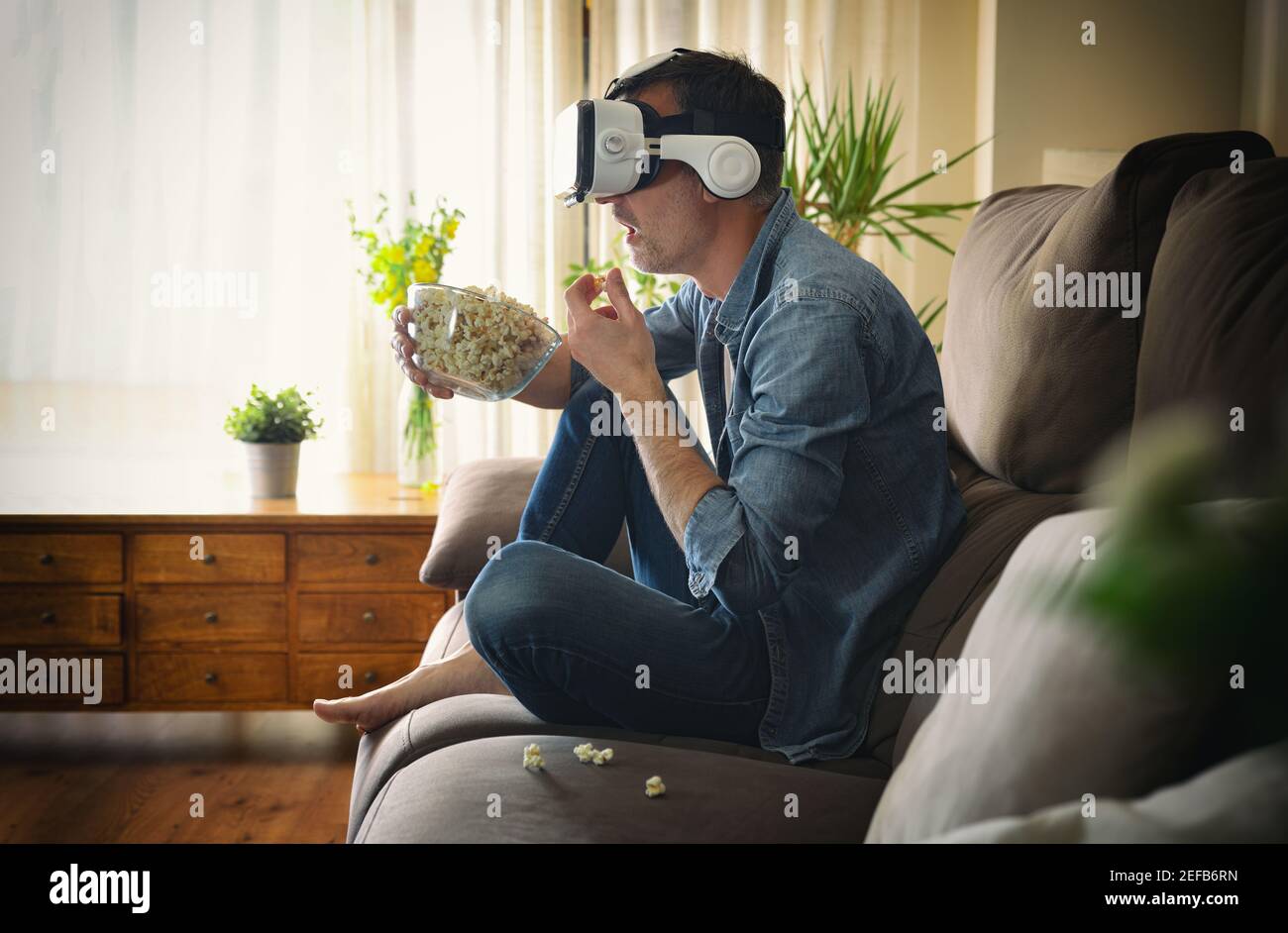 Un uomo stupefatto che guarda un film in occhiali di realtà virtuale e. mangiare popcorn seduto sul divano a casa Foto Stock