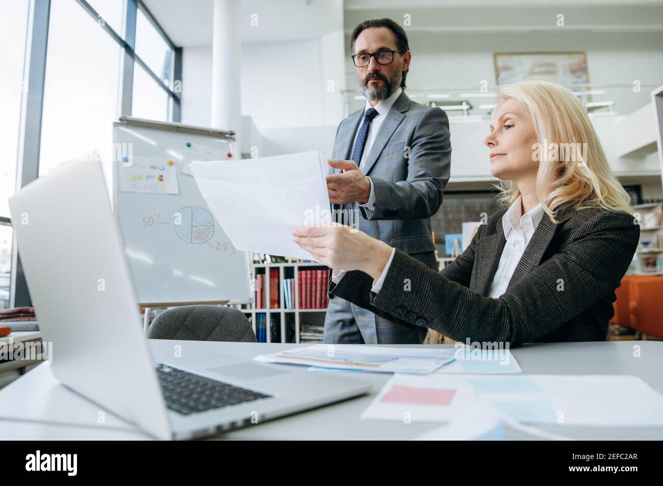 Il maschio e la femmina maturi seri stanno imparando i grafici finanziari, facendo i documenti all'ufficio. Bella donna bionda e attraente uomo che lavorano insieme su un importante progetto di business Foto Stock