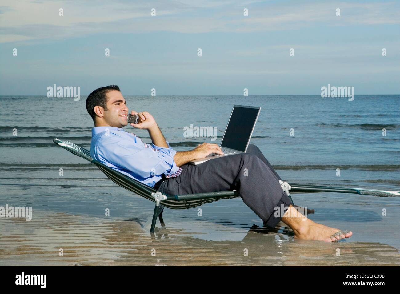 Profilo laterale di un giovane seduto su un salotto sedia sulla spiaggia che parla su un telefono cellulare e. utilizzando un computer portatile Foto Stock