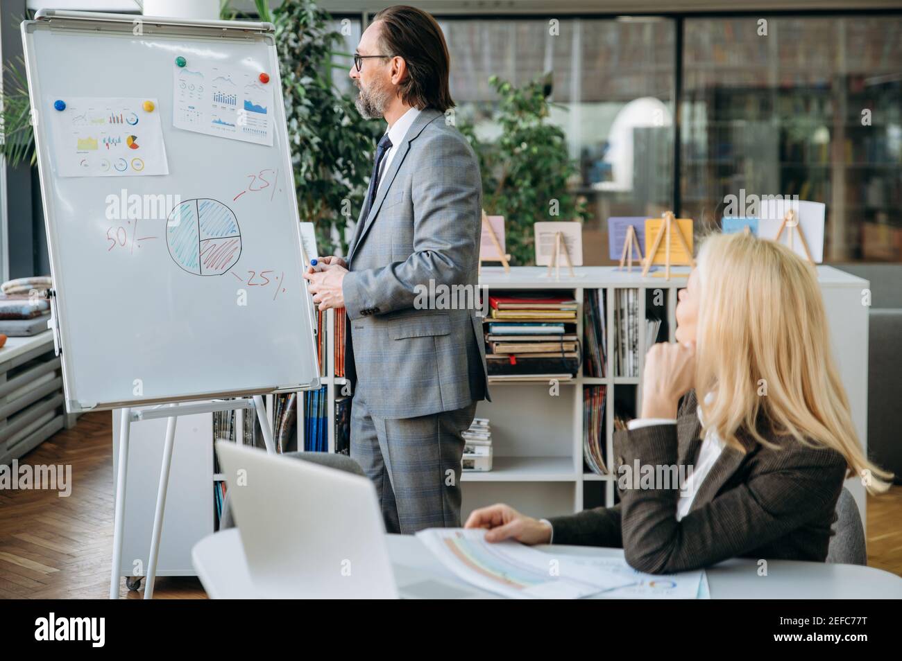 Un elegante dipendente di mezza età invia un rapporto finanziario, facendo una presentazione e mostrando grafici sulla bacheca. Un uomo elegante sta in piedi vicino alla lavagna a fogli mobili in ufficio, discutendo il piano di lavoro Foto Stock