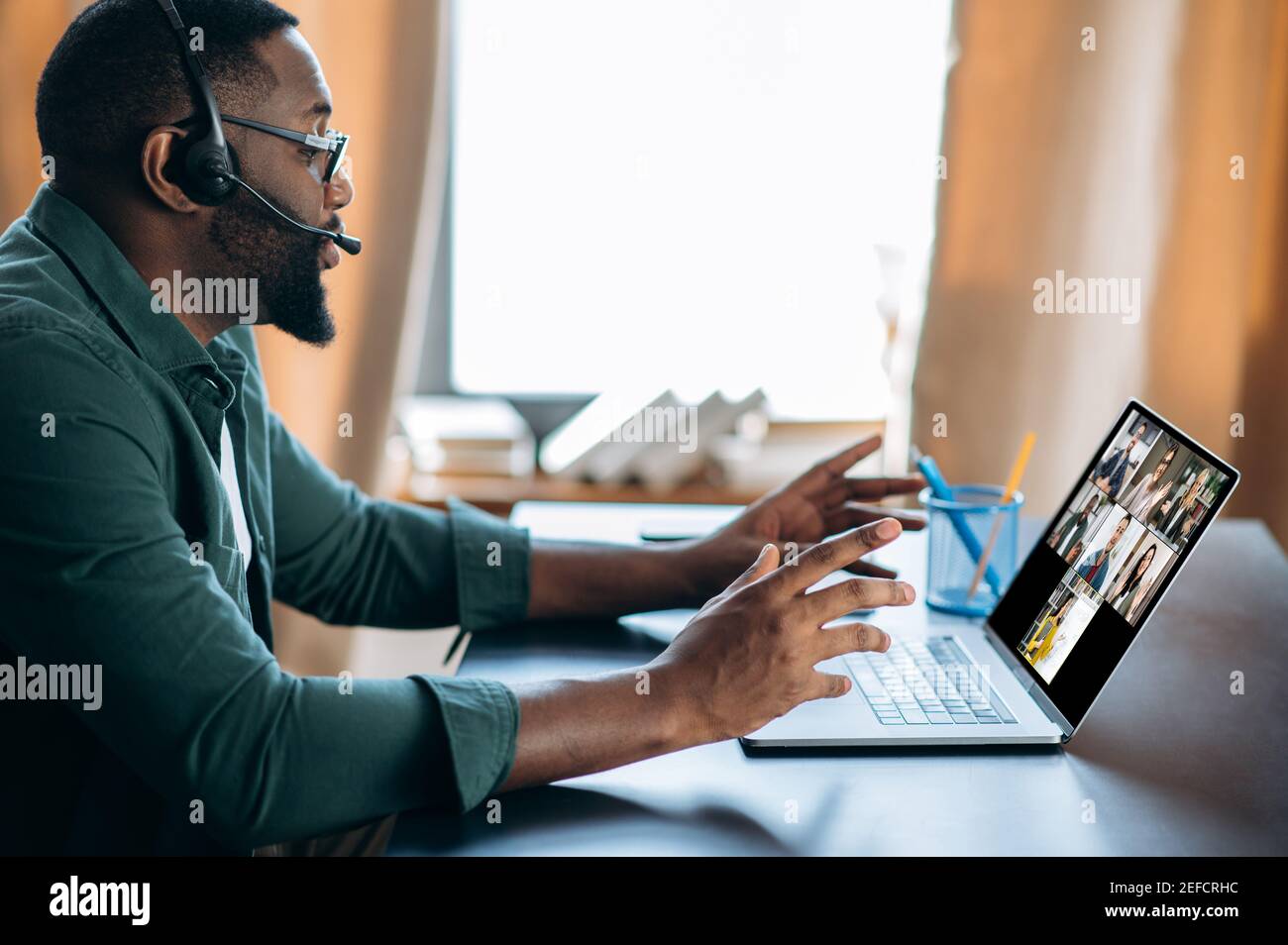 Conferenza virtuale con persone multietniche. Un ragazzo afro-americano in cuffia impara a distanza utilizzando il laptop e l'app di chat video. Sullo schermo del portatile l'insegnante e il gruppo di persone diverse, online Foto Stock