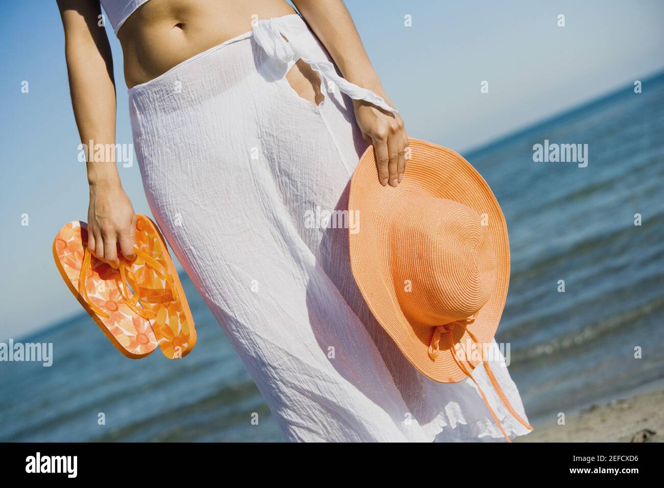 Vista in sezione intermedia di una donna che tiene il cappello e un coppia di flip flop sulla spiaggia Foto Stock