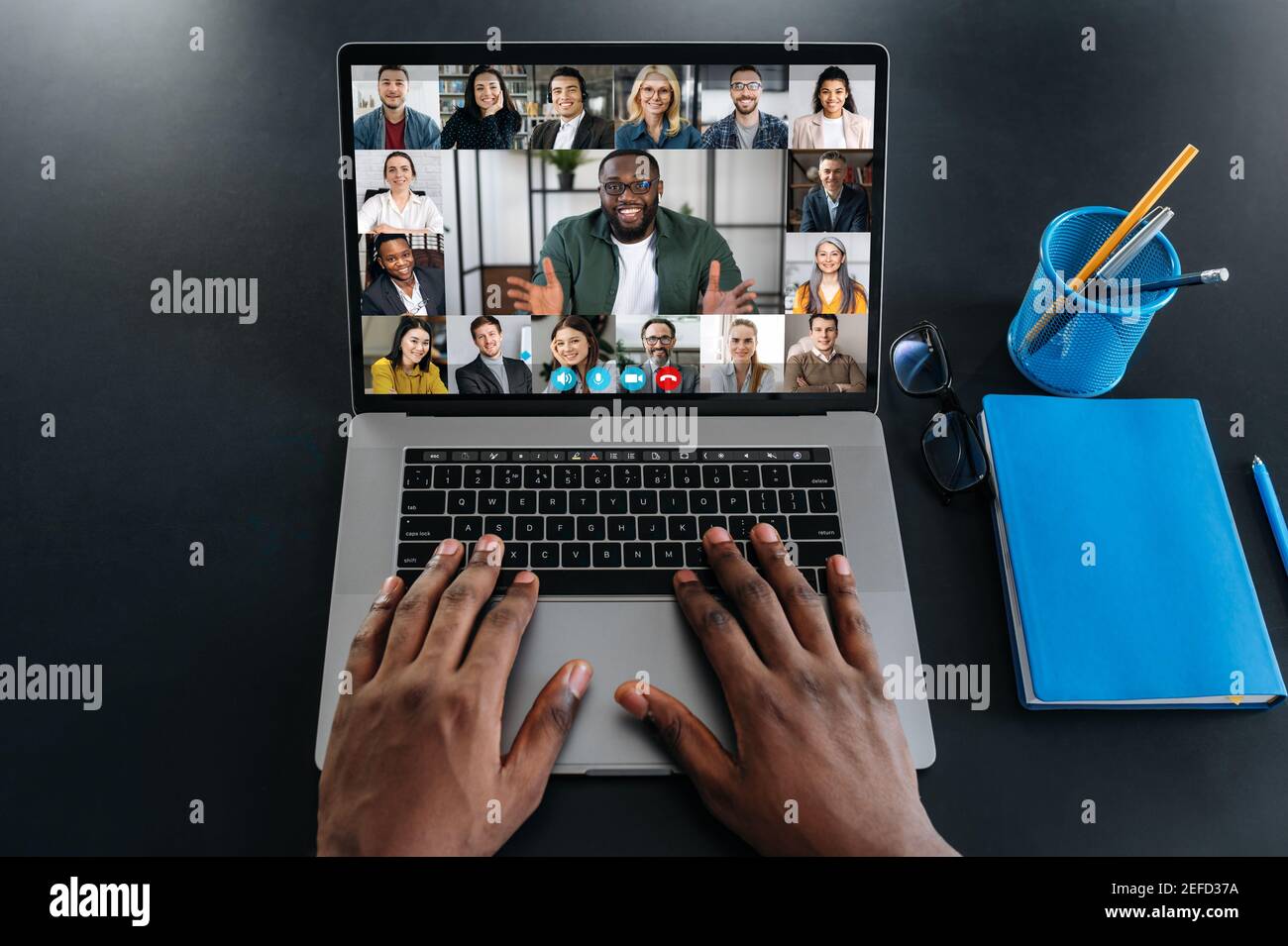 Riunione di affari in linea. Uomini d'affari multirazziali che lavorano da casa con una videoconferenza. Vista dall'alto sullo schermo di un computer portatile con webcam e immagini di persone diverse e mani di un uomo nero su una tastiera Foto Stock
