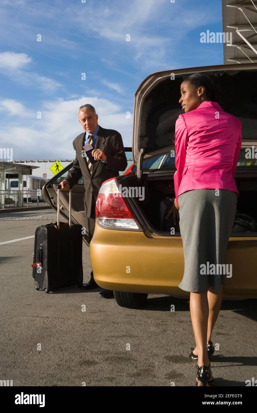 Donna d'affari che scarica la sua valigia da un bagagliaio auto con un uomo d'affari in piedi davanti a lei Foto Stock