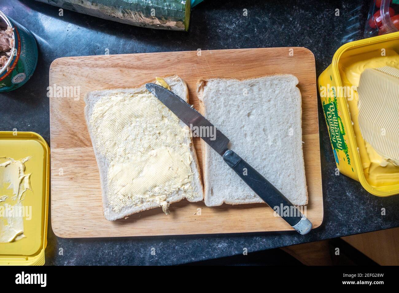 Pane bianco su un tagliere di legno e una vasca di burreria spalmata pronta per iniziare a preparare un panino per il pranzo. Foto Stock