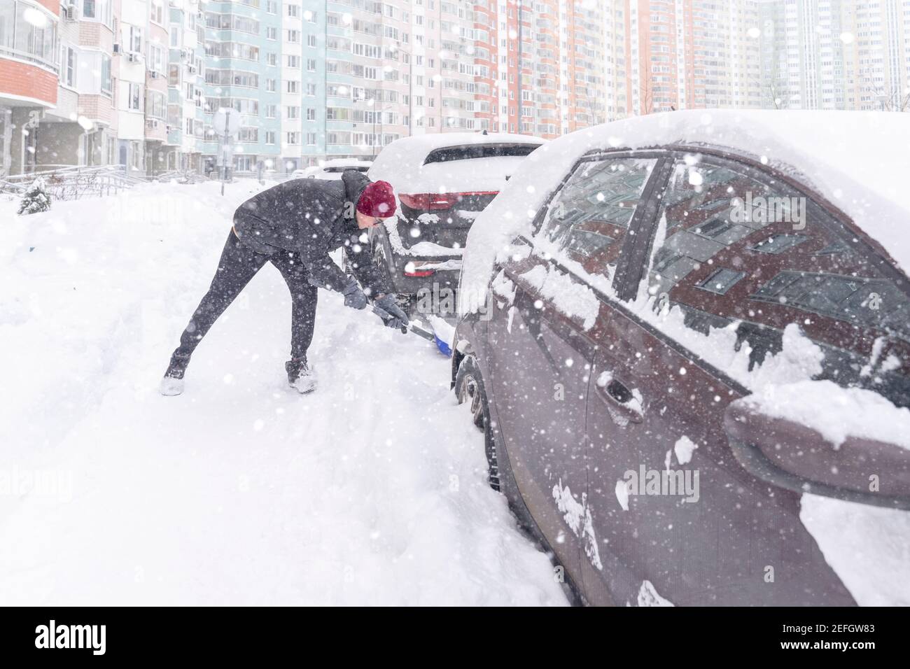 un giovane in una giacca calda e un cappello sta scavando la sua auto dalla cattività della neve con una pala. nevicata pesante, scarsa visibilità Foto Stock