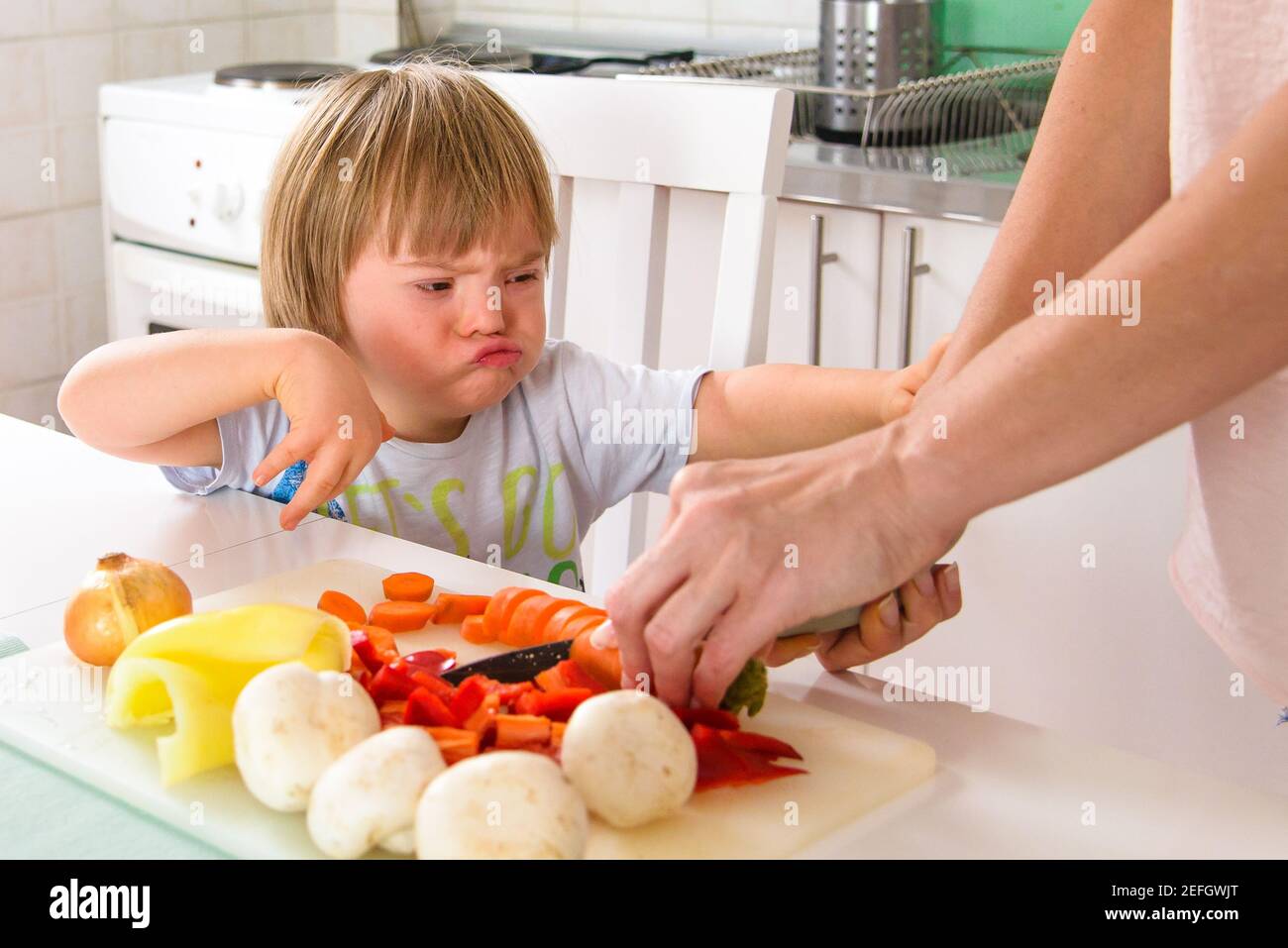 Adorabile ragazzo sindrome Down mostrando insoddisfazione e frustrazione con sano preparazione del cibo da parte della madre durante la quarantena Foto Stock
