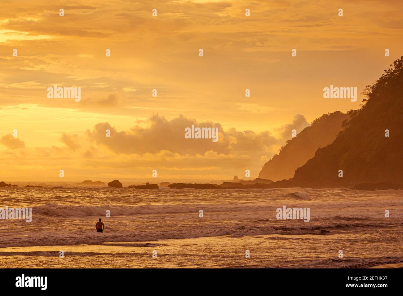 Silhouette di uomo irriconoscibile, all'interno dell'acqua guardando il tramonto sulla riva. Spiaggia di Jaco nel Pacifico della Costa Rica. Foto Stock