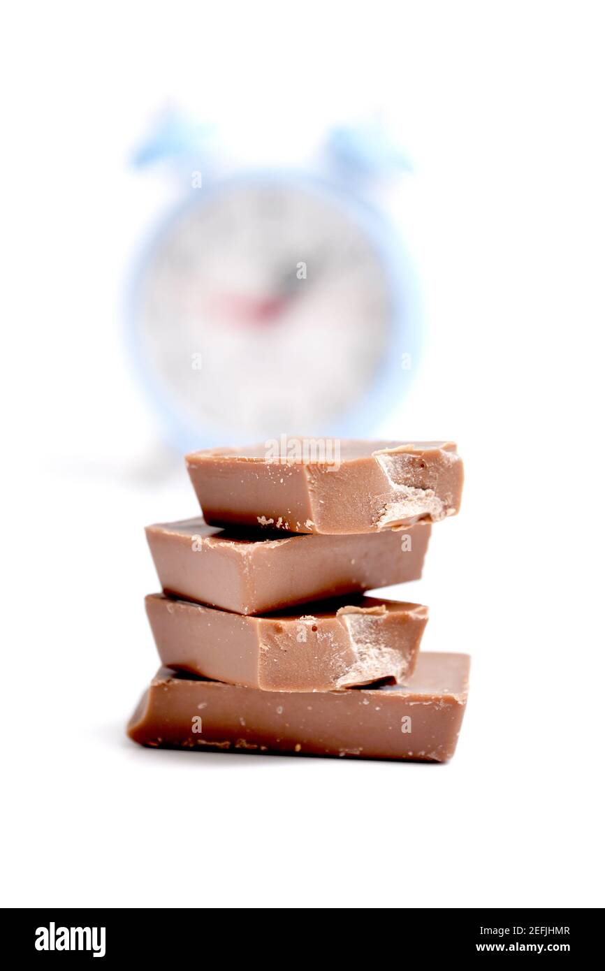 Le barrette di cioccolato si impilano davanti alla sveglia blu sfocata su sfondo bianco Foto Stock